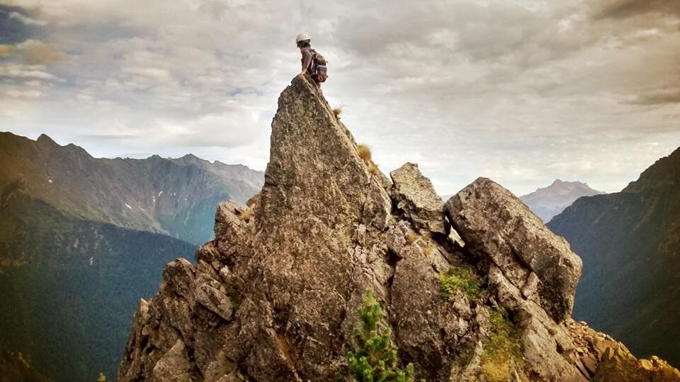 Fabien, a friend of Anthony, the owner of the website, sitting at the top of a tall pointy stone, facing away from the camera, on the top of a mountain, in the French Alps