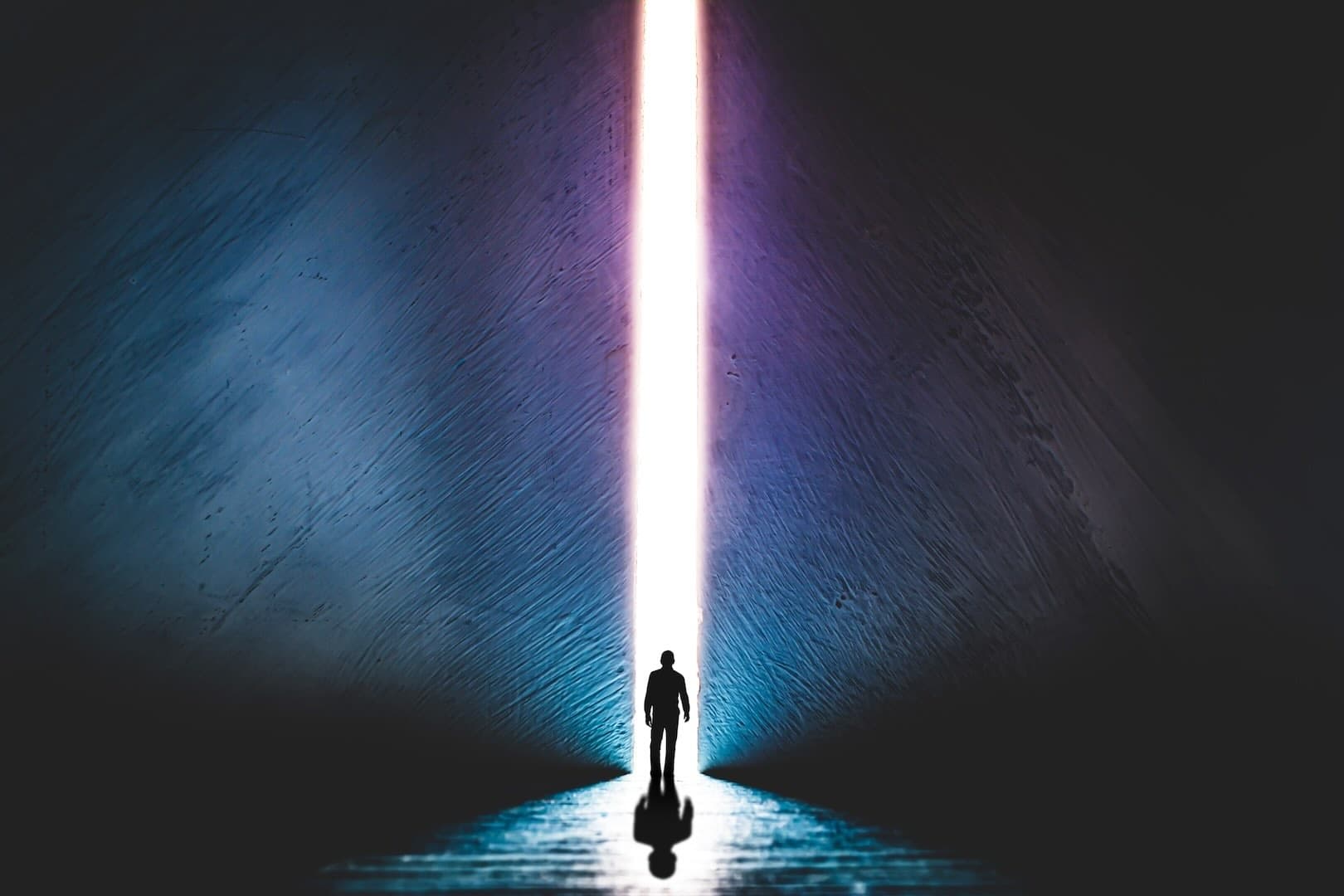 Silhouette of a person standing in front tall ray of light created by the opening between 2 big walls, looking like he is about to enter the light