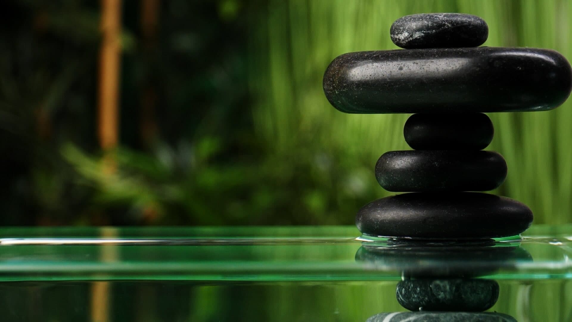 A few flat black stones stacked on top of each other, with a drop of water dropping into the water, creating a ripple