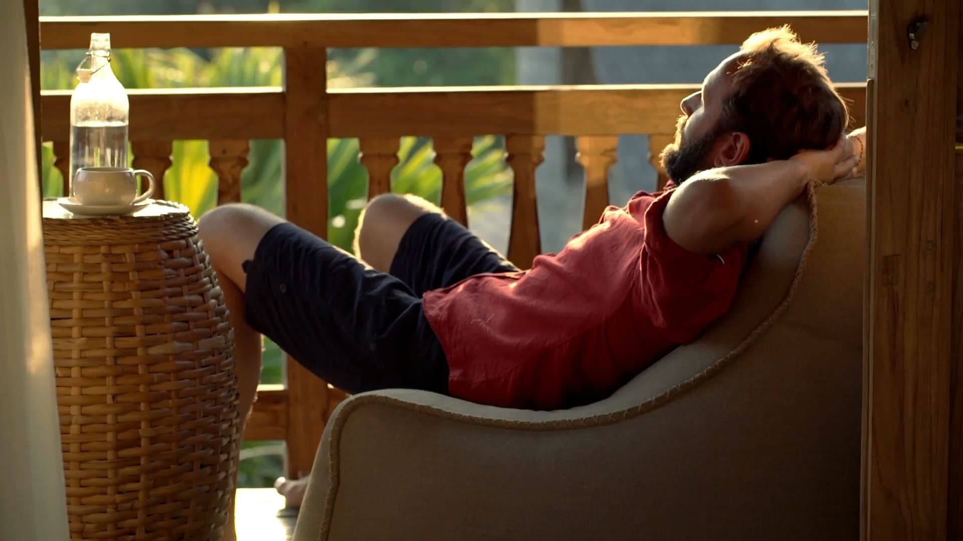 A man laying back in a lounge chair on a terrace with nature in the background, relaxing and putting a hat over his face, to relax