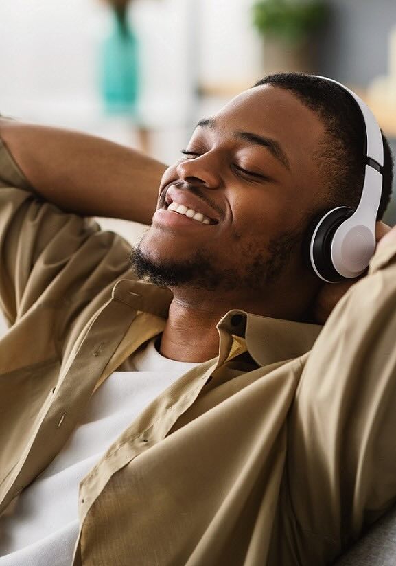 Black male wearing white headphones and eyes closed, relaxing on a couch, with his hands behind his head