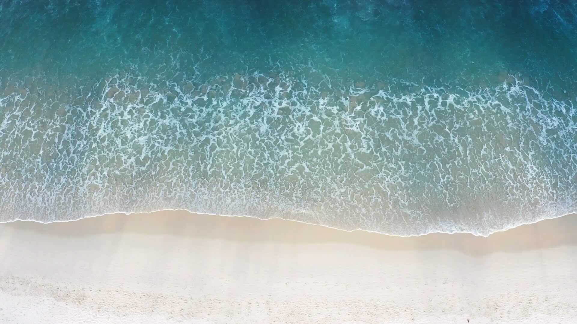 A view from above, of a beach, with waves of a deep blue water gently crashing on the sand