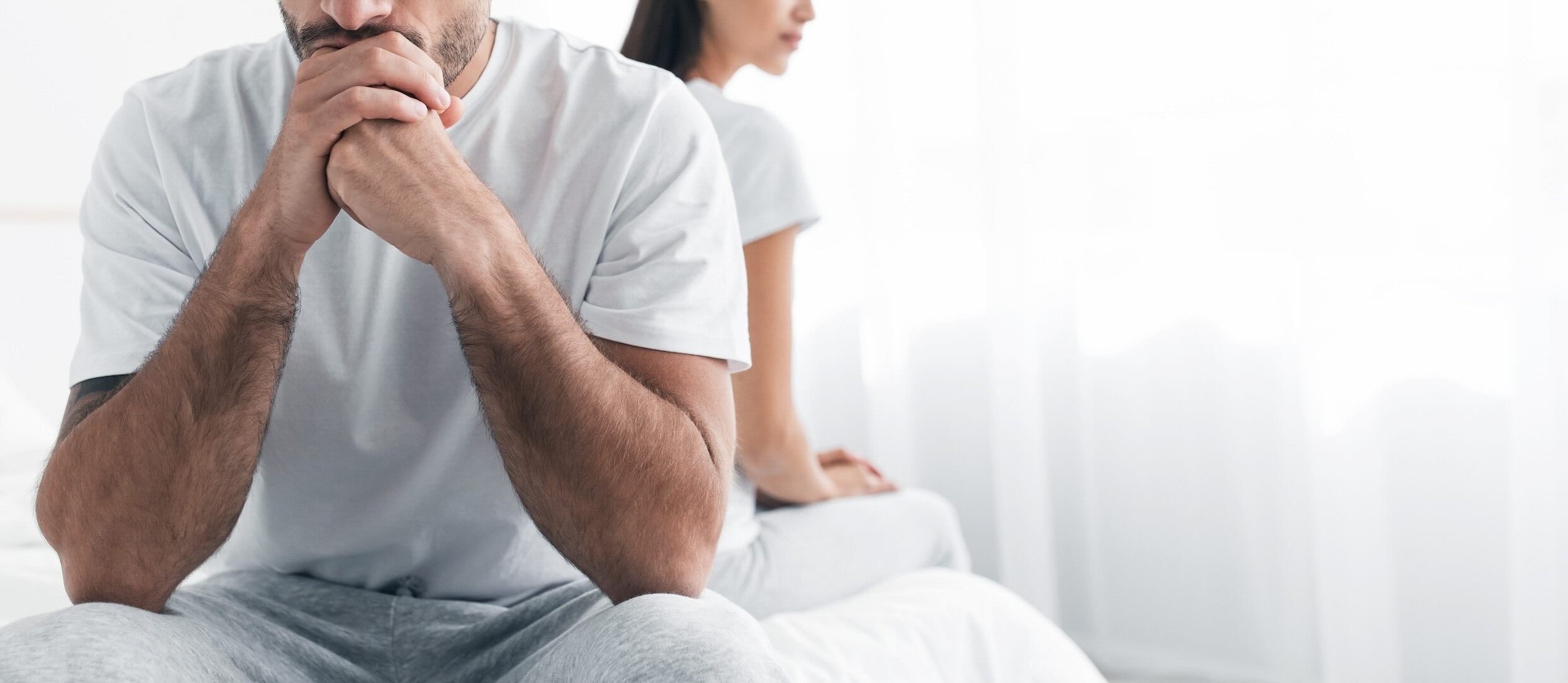 A white male facing the camera and a white female behind him, sitting back-to-back on the edges of a bed, in an apparently tensed situation