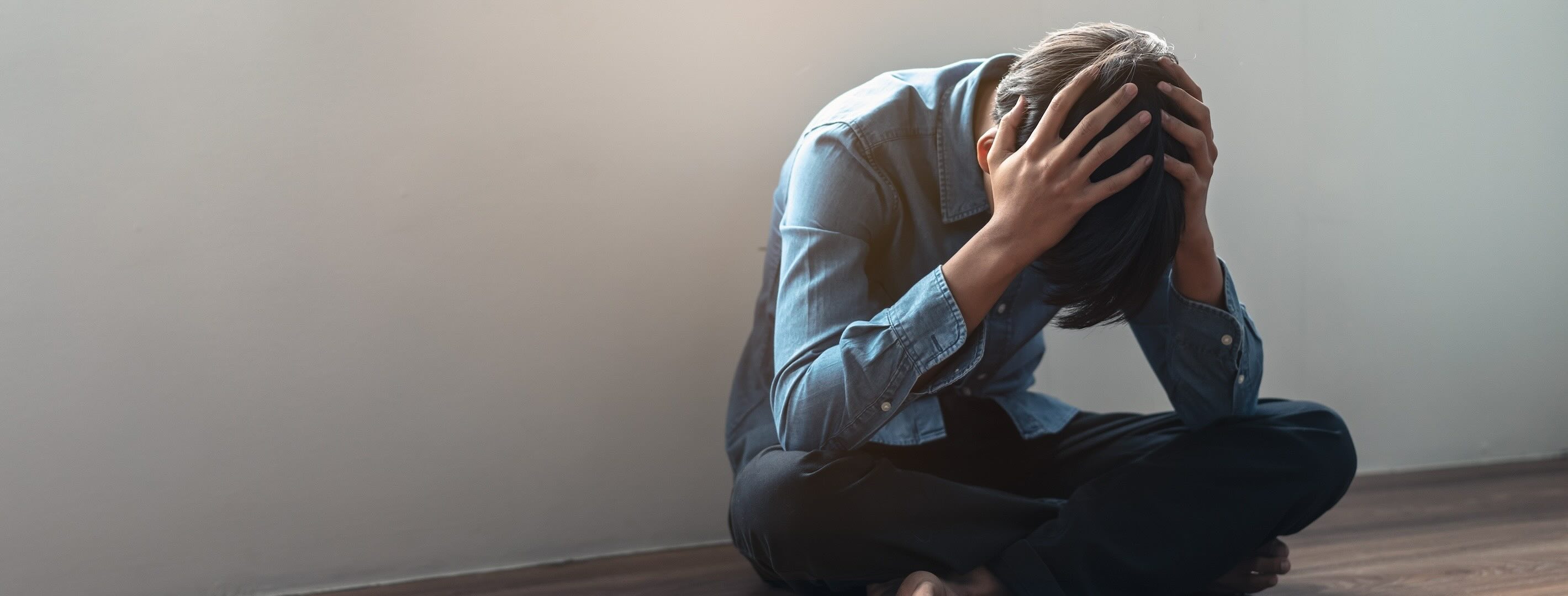 Young white male sitting cross-legged on the floor, with his elbows on his legs, holding his head in his hands, in apparent distress